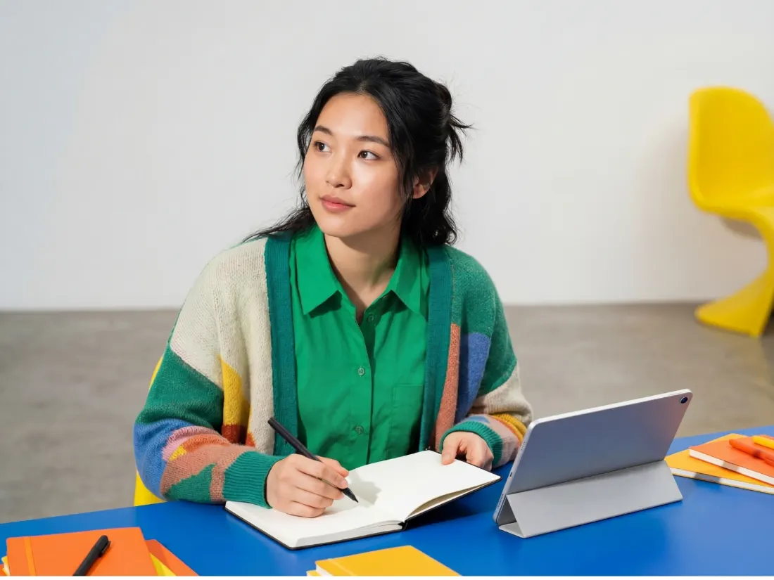 A woman pausing mid-thought at a desk with a notebook and tablet